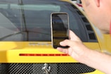 A close-up shot of a person holding a smartphone in front of a yellow sports car. The phone screen displays a photo being taken, which includes part of the car's rear. The car features a red brake light strip, black mesh near the exhaust area, and notable badging.