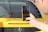 A close-up shot of a person holding a smartphone in front of a yellow sports car. The phone screen displays a photo being taken, which includes part of the car's rear. The car features a red brake light strip, black mesh near the exhaust area, and notable badging.