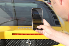 A close-up shot of a person holding a smartphone in front of a yellow sports car. The phone screen displays a photo being taken, which includes part of the car's rear. The car features a red brake light strip, black mesh near the exhaust area, and notable badging.