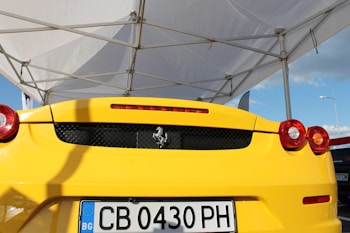 A yellow sports car is parked under a white canopy with a visible Ferrari logo on the rear. The car's sleek and glossy design is accentuated by the bright sunlight, highlighting the tail lights and rear features. A license plate from Bulgaria is prominently displayed.