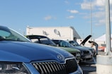 A parking lot scene with several cars parked, including a blue car prominently in the foreground. Two people are inspecting a car with its hood open in the background. The sky is clear with a few clouds, and a white building with satellite dishes is visible.