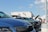 A parking lot scene with several cars parked, including a blue car prominently in the foreground. Two people are inspecting a car with its hood open in the background. The sky is clear with a few clouds, and a white building with satellite dishes is visible.