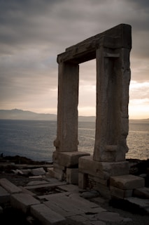 A large, ancient stone gateway stands prominently against a backdrop of a vast sea and distant mountains under a cloudy, overcast sky. The structure appears weathered, suggesting historical significance.