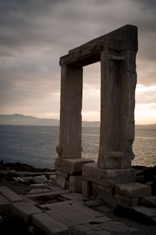 A large, ancient stone gateway stands prominently against a backdrop of a vast sea and distant mountains under a cloudy, overcast sky. The structure appears weathered, suggesting historical significance.