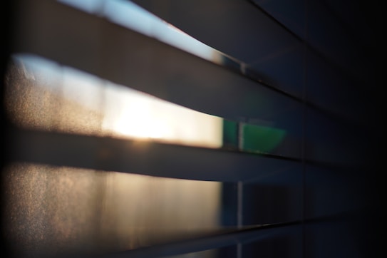 Horizontal slats of window blinds partially open, allowing light to filter through, creating a soft, diffused glow with shadows and highlights.