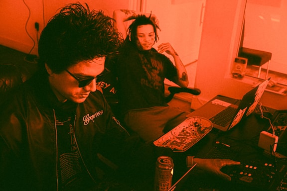 Two individuals are indoors, engaged with what appears to be music production equipment. The person on the left is wearing sunglasses and a jacket with '999' embroidered on it, focused on operating a mixer. The person on the right, sitting back with a relaxed posture, has a skateboard on their lap and a laptop in front of them. The lighting is heavily tinted in red, creating a warm ambiance.
