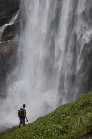A person with a backpack stands at the edge of a lush, green hill, gazing at a massive waterfall cascading down the rocky cliffside. The mist from the waterfall fills the air, creating a sense of power and grandeur.
