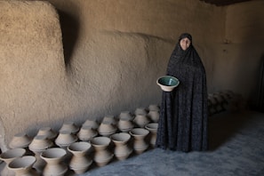 A person stands in a dimly lit, rustic room holding a ceramic pot with a green interior. The walls are made of plaster or clay, and the space is lined with numerous similar pottery pieces on the floor.