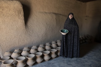 A person stands in a dimly lit, rustic room holding a ceramic pot with a green interior. The walls are made of plaster or clay, and the space is lined with numerous similar pottery pieces on the floor.