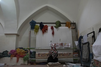 A person wearing a patterned headscarf is sitting in front of a traditional loom, working on a colorful tapestry. The room has vaulted ceilings, and multiple bundles of yarn in various colors are hanging above and scattered around. The tapestry in progress features intricate designs and a variety of colors.