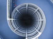 An architectural structure featuring a circular, multi-level, spiral concrete ramp leading into a dark central core. Surrounding the structure are blue walls, and a few red traffic cones are visible around the edges.