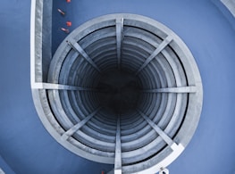 An architectural structure featuring a circular, multi-level, spiral concrete ramp leading into a dark central core. Surrounding the structure are blue walls, and a few red traffic cones are visible around the edges.