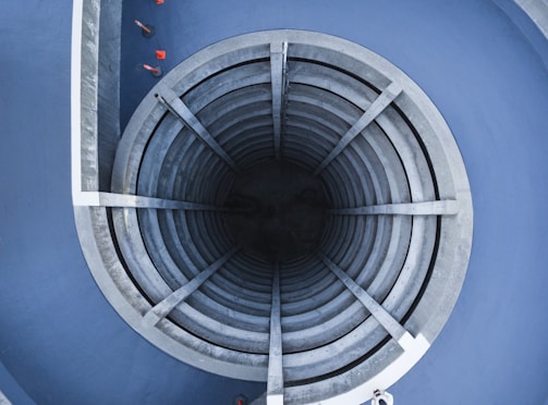 An architectural structure featuring a circular, multi-level, spiral concrete ramp leading into a dark central core. Surrounding the structure are blue walls, and a few red traffic cones are visible around the edges.