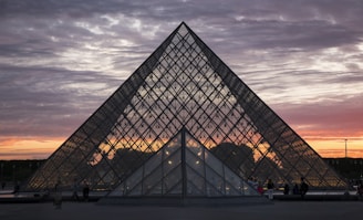 A glass pyramid structure set against a dramatic sunset sky, with clouds dispersed in hues of purple, pink, and orange. Soft lighting illuminates from within the pyramid, and silhouettes of people are visible near its base.