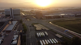 An aerial view of an industrial and commercial area with large buildings and expansive parking lots, set against a serene sunrise or sunset. The bright, warm sun casts a golden glow across the landscape. There are several trucks parked in rows, and the area is surrounded by roads and patches of greenery.