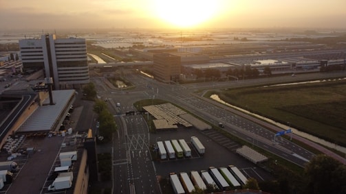 An aerial view of an industrial and commercial area with large buildings and expansive parking lots, set against a serene sunrise or sunset. The bright, warm sun casts a golden glow across the landscape. There are several trucks parked in rows, and the area is surrounded by roads and patches of greenery.