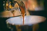 A close-up view of a coffee machine spout pouring a rich, golden-brown espresso shot into a white cup, with a blurred background creating a warm and cozy atmosphere.