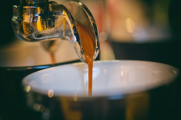 A close-up of a steaming espresso shot with golden crema, set against a dark charcoal background with soft gold light accents.