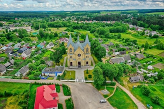 An aerial view of a village features a prominent church with twin spires in the center, surrounded by lush greenery and small houses. The area includes well-maintained gardens and roads, with a vibrant mix of natural and architectural elements.