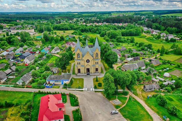 An aerial view of a village features a prominent church with twin spires in the center, surrounded by lush greenery and small houses. The area includes well-maintained gardens and roads, with a vibrant mix of natural and architectural elements.