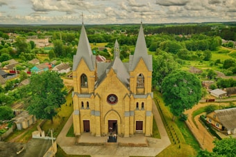 A large, ornate brick church with pointed arches and three tall spires stands prominently in a rural area. Surrounding the church is a lush, green landscape with trees and scattered houses. The sky above is partly cloudy, adding depth to the vibrant scenery.