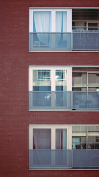 Three levels of a modern apartment building are visible, featuring large glass windows and metal balconies. Each balcony has a mesh railing, and the windows are adorned with curtains. The building's facade is composed of red brick.