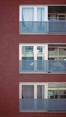 Three levels of a modern apartment building are visible, featuring large glass windows and metal balconies. Each balcony has a mesh railing, and the windows are adorned with curtains. The building's facade is composed of red brick.