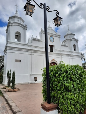 A historic white church with two bell towers and a blue sky above. A black iron lamppost stands in front, surrounded by green foliage. The architecture features intricate details with crosses atop each spire. The atmosphere feels tranquil and serene.