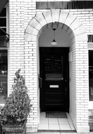 An arched entrance framed by light-colored bricks leads to a dark wooden door. A lamp hangs from the center of the arch. There is a potted plant on the left side and a welcome mat on the tiled floor in front of the door.
