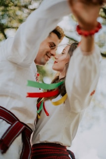 A couple dressed in traditional clothing shares an intimate moment, with ribbons in various colors attached to their garments. The background is blurred, highlighting the focus on the couple and creating a warm, joyful atmosphere.
