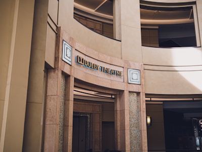 An entrance to a building labeled 'Dolby Theatre' with architectural features including rounded walls and columns. The signage has modern lettering, and there are decorative mosaic patterns flanking the entrance.