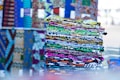 A stack of colorful, patterned fabrics neatly folded and stacked on a reflective surface, creating an intricate display of vibrant geometric and abstract designs. The fabrics are likely arranged for sale in a market or shop, with a variety of patterns incorporating colors like blue, yellow, green, purple, and orange. In the background, additional rolls or stacks of similar fabrics can be seen, each showcasing unique and bold patterns.