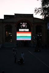 An industrial-style brick building with a large arched window displaying a colorful neon sign. The sign features the word 'SCHIFFBAU' and a graphic with a rainbow banner stating 'Love is Love.' There are several people in the foreground, including someone in a wheelchair.