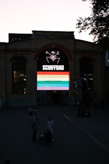 An industrial-style brick building with a large arched window displaying a colorful neon sign. The sign features the word 'SCHIFFBAU' and a graphic with a rainbow banner stating 'Love is Love.' There are several people in the foreground, including someone in a wheelchair.