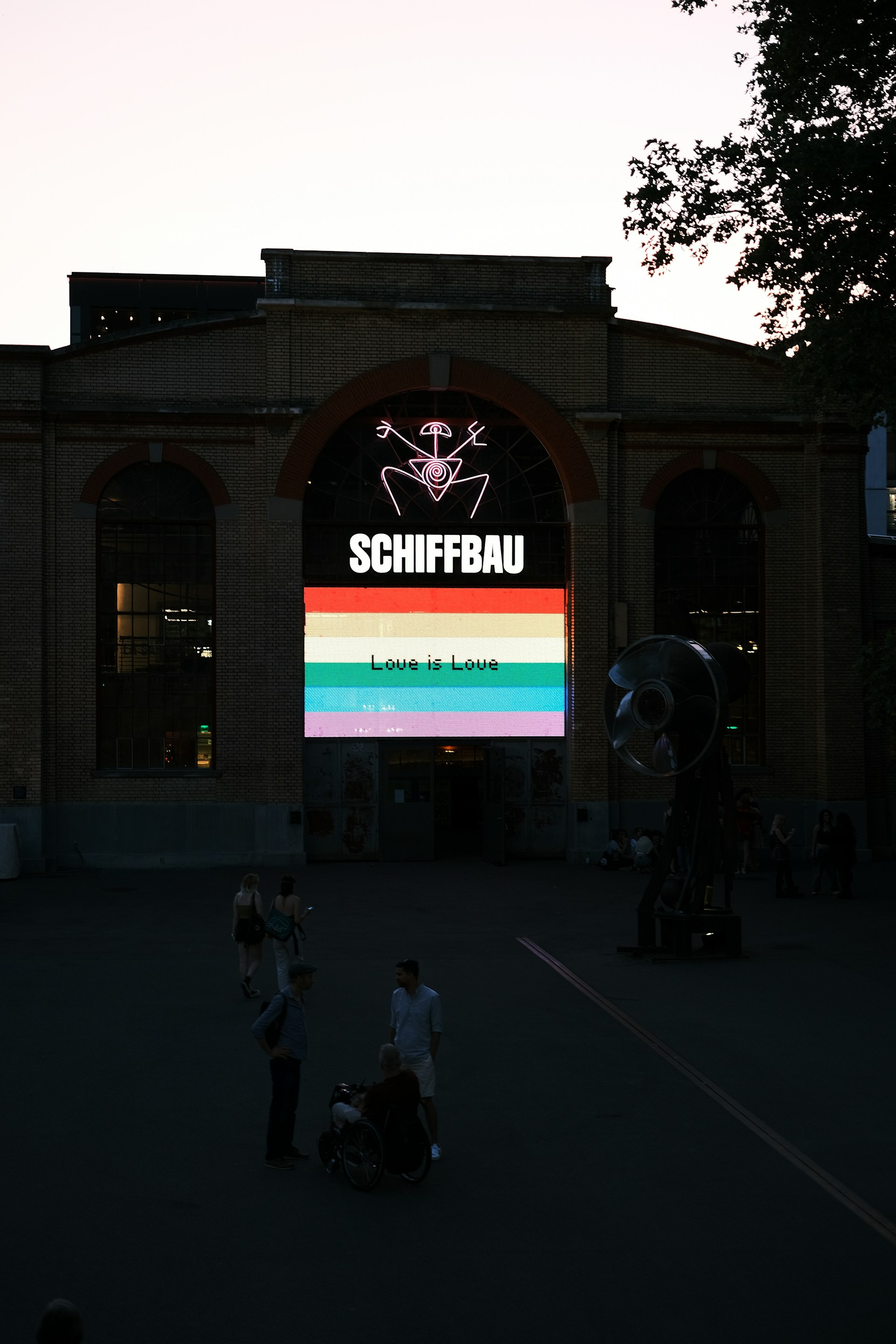 An industrial-style brick building with a large arched window displaying a colorful neon sign. The sign features the word 'SCHIFFBAU' and a graphic with a rainbow banner stating 'Love is Love.' There are several people in the foreground, including someone in a wheelchair.