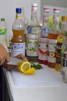A person is slicing a lemon on a cutting board. Various bottles of flavored syrup are arranged on the countertop, including banana, wild mint, watermelon, peach, and apricot flavors. Fresh mint leaves are also present alongside the lemon slices.