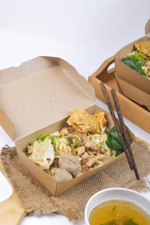 A serving of noodles with meatballs, vegetables, and fried wontons is presented in a brown cardboard takeout box. Two dark-colored chopsticks rest on the edge of the box, and a bowl of clear soup is placed next to it on a woven mat. The setting has a clean and simple layout with natural textures.