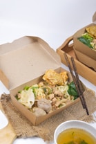 A serving of noodles with meatballs, vegetables, and fried wontons is presented in a brown cardboard takeout box. Two dark-colored chopsticks rest on the edge of the box, and a bowl of clear soup is placed next to it on a woven mat. The setting has a clean and simple layout with natural textures.