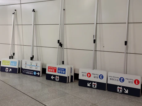 A row of portable signs with telescopic poles, leaning against a tiled wall. Each sign displays information in multiple languages, including arrows and symbols likely related to public transportation routes or stations.