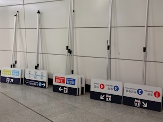 A row of portable signs with telescopic poles, leaning against a tiled wall. Each sign displays information in multiple languages, including arrows and symbols likely related to public transportation routes or stations.