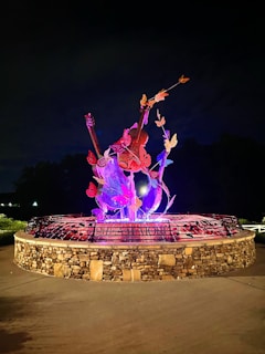 A vibrant sculpture featuring colorful butterflies surrounding musical instruments, illuminated against a dark background. The base is made of stone, adding texture and contrast to the vivid colors.
