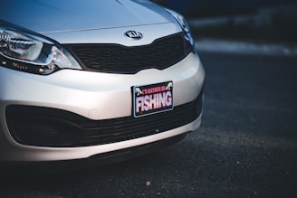 A close-up view of the front portion of a silver Kia car, focusing on the license plate area. The license plate frame features a sign with the text 'I'D RATHER BE FISHING' written in bold letters.