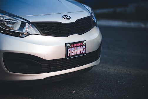 A close-up view of the front portion of a silver Kia car, focusing on the license plate area. The license plate frame features a sign with the text 'I'D RATHER BE FISHING' written in bold letters.