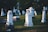 A serene graveyard with several white tombstones set against a backdrop of green grass and trees. The stones vary in height and design, with some having crosses on top. Soft sunlight casts gentle shadows, creating a peaceful and solemn atmosphere.