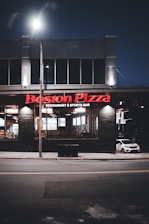A Boston Pizza restaurant and sports bar at night. The building features large windows through which the interior lights are visible, creating a welcoming glow. The signage is prominently displayed in red above the entrance. A car is parked on the side, and the scene is illuminated by streetlights.