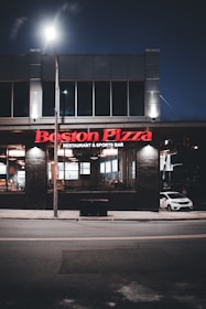 A Boston Pizza restaurant and sports bar at night. The building features large windows through which the interior lights are visible, creating a welcoming glow. The signage is prominently displayed in red above the entrance. A car is parked on the side, and the scene is illuminated by streetlights.