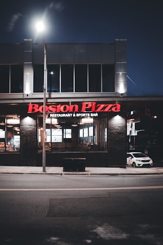 A Boston Pizza restaurant and sports bar at night. The building features large windows through which the interior lights are visible, creating a welcoming glow. The signage is prominently displayed in red above the entrance. A car is parked on the side, and the scene is illuminated by streetlights.