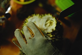 A gloved hand is holding a durian on a wooden surface. A knife is positioned near the durian, suggesting it is about to be cut open. The scene is dimly lit, with shadows creating a focused and slightly mysterious atmosphere.