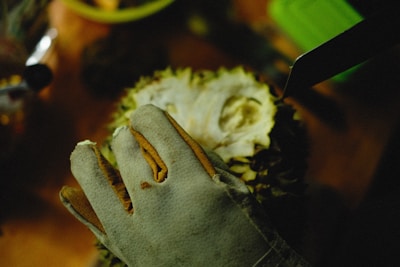 A gloved hand is holding a durian on a wooden surface. A knife is positioned near the durian, suggesting it is about to be cut open. The scene is dimly lit, with shadows creating a focused and slightly mysterious atmosphere.