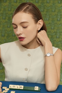 A young woman with short, slicked-back hair is dressed in a sleeveless, white top adorned with pearl-like buttons. She rests her chin on her hand, which bears an elegant wristwatch. In front of her on a blue surface lies a row of letter tiles spelling out 'LOLA', along with an additional tile featuring the letter 'E'. The backdrop is a blurred, textured green pattern.